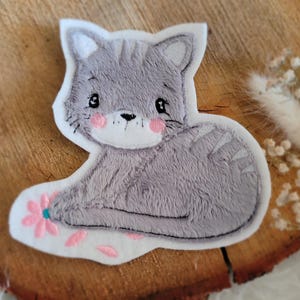 Könnte beinhalten: Ein grauer Plüschkatze mit rosa Wangen und einem weißen Umriss, die auf einem weißen Hintergrund mit rosa Blumen sitzt.