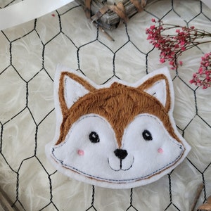 Könnte beinhalten: Ein weißer und brauner Plüsch-Fuchs-Applik mit gestickten Augen, Nase und Mund. Der Fuchs lächelt und hat rosa Wangen.