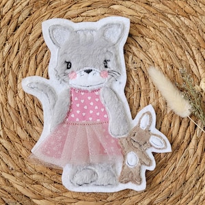 Könnte beinhalten: Ein graues, katzenförmiges Filz-Applikation, das ein rosa Kleid mit weißen Herzen und einen glitzernden Tüllrock trägt. Die Katze hält eine kleine braune Hasen-Applikation. Der Hintergrund ist eine gewebte, hellbraune Oberfläche.
