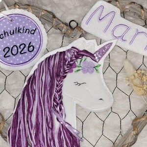 Könnte beinhalten: Ein herzförmiger Drahtrahmen mit einem weißen Einhorn-Applikation mit lila Mähne und glitzerndem Horn. Enthält ein Etikett mit "Schulkind 2026" und ein Schild mit "Marie".