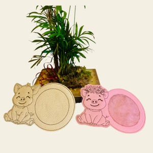 Könnte beinhalten: Zwei Untersetzer in Schweineform, einer mit einer Blumenkrone und der andere mit einem leeren Platz für Personalisierung. Die Untersetzer sind aus weichem, rosa Material und haben eine leicht strukturierte Oberfläche.