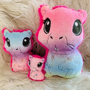 May include: Three plush toys in the shape of a cartoon animal with large eyes and a pink, blue, and purple ombre color scheme. The largest toy is pink and the smallest toy is pink. The middle toy is blue.