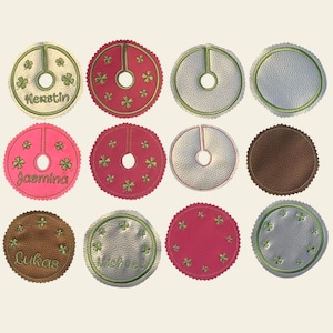 Puede incluir: Doce faldas de árbol de Navidad redondas de fieltro en varios colores, incluyendo marrón, rosa, rojo y verde. Cada falda tiene un agujero en el centro para el tronco del árbol y está decorada con tréboles verdes y un nombre bordado en hilo verde. Los nombres incluyen "Kerstin", "Jasmina", "Lukas" y "Michael".