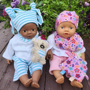 Op de afbeelding: Twee babypoppen in zachte kleding. Eén pop draagt een blauw-wit gestreepte hoed en broek en houdt een kleine teddybeer vast. De andere pop draagt een roze bloemenhoed en slabbetje en houdt een roze bloemendeken vast.