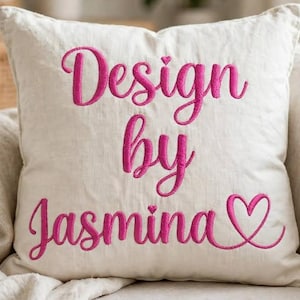 Könnte beinhalten: Ein quadratisches, cremefarbenes Zierkissen mit dem gestickten Text "Design by Jasmina" in einer leuchtend pinkfarbenen Schreibschrift. Das Design enthält ein kleines Herz über dem Buchstaben i und ein größeres Herzsymbol am Ende des Namens.