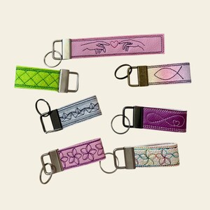 May include: A collection of embroidered keychains in various colors and designs. The keychains feature different patterns, including hands with a heart, a fish symbol, geometric shapes, and floral motifs. Each keychain has a metal clasp and ring.