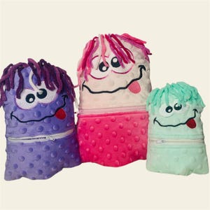May include: Three plush monster toys with embroidered faces and fuzzy textures. The monster on the left is purple, the middle monster is pink, and the monster on the right is green.