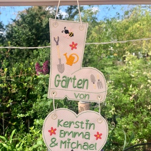 May include: A white fabric garden sign with a green boot shape and a heart shape. The boot has a bee, a watering can, a shovel, and the text "Garten von". The heart has a bee, two red flowers, and the text "Kerstin Emma & Michael".