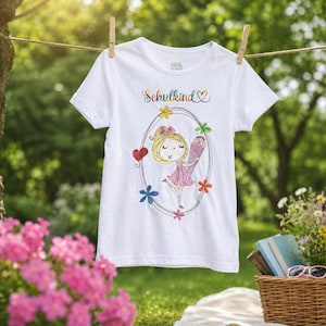 Puede incluir: Una camiseta blanca de manga corta con un gráfico bordado de colores que muestra a una niña sosteniendo un cono, rodeada por un marco ovalado y flores. El texto "Schulkind" está impreso en degradado arcoíris sobre la ilustración.