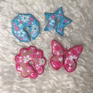 以下が含まれることがあります： 星、八角形、花、蝶の形をした4つの布製アップリケ。各アップリケは、青またはピンクの背景にピンクと白の花柄があります。それぞれにボタン留めが付いています。