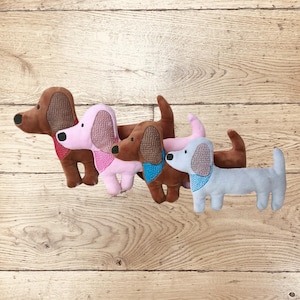 Könnte beinhalten: Vier Plüsch-Dackel-Spielzeuge in den Farben Braun, Rosa und Hellblau. Jeder Hund hat einen andersfarbigen Schal und Ohrendetails. Die Spielzeuge sind auf einer Holzoberfläche angeordnet.