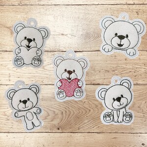 Könnte beinhalten: Fünf gestickte Teddybär-Ornamente in verschiedenen Posen. Die Bären sind hellgrau mit schwarzen und braunen Details. Ein Bär hält ein rotes Herz. Jedes Ornament hat oben eine kleine Schlaufe zum Aufhängen.