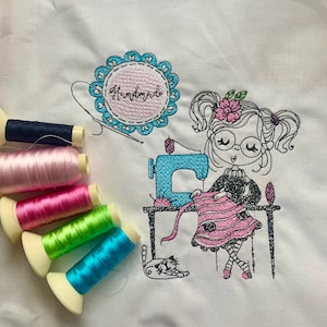 Könnte beinhalten: Stickerei mit einer Mädchenfigur mit einer Nähmaschine, die ein Stück Stoff hält. Das Design enthält das Wort "Handmade" in einem Kreis und eine Katze. Das Design ist auf einem weißen Stoffhintergrund.