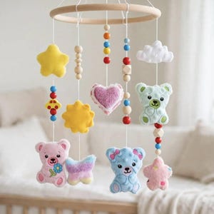 May include: A nursery mobile featuring a circular wooden frame with hanging strings of colorful wooden beads and plush felt shapes. The hanging elements include yellow stars, a white cloud, a purple heart, a yellow sun, and light blue and pink teddy bears.