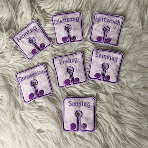 May include: Set of seven embroidered fabric garment dividers, each labeled with a day of the week in purple thread. The dividers have a floral pattern and a keyhole shape with a button detail. The days of the week are: Monday, Tuesday, Wednesday, Thursday, Friday, Saturday, and Sunday.