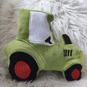 Puede incluir: Un peluche verde con forma de tractor con ruedas negras y centro rojo. El tractor tiene una ventana blanca y detalles negros. El juguete está hecho de tela suave y texturizada y está diseñado para niños.