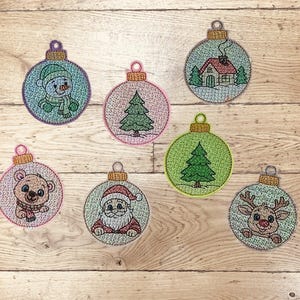 Könnte beinhalten: Gestickte Weihnachtskugeln mit verschiedenen Designs, darunter ein Schneemann, Weihnachtsbaum, Haus, Bär, Weihnachtsmann und Rentier. Jede Kugel ist rund mit einem metallisch goldenen Aufsatz und einer Schlaufe zum Aufhängen. Die Farben sind Blau, Rosa und Grün.