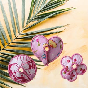 Könnte beinhalten: Drei rosa Stilleinlagen in verschiedenen Formen: ein Herz, eine Blume und ein Kreis. Jedes Pad hat eine Schlaufe und einen Knopfverschluss. Die Herz- und Blumen-Pads haben florale Muster, während das Kreis-Pad ein gemustertes Design hat.
