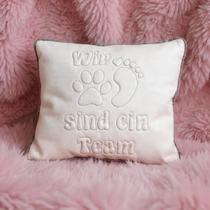 May include: A square, light pink pillow with a gray border. The pillow features embroidered text and designs, including a paw print, a footprint, and the words "Wir sind ein Team."