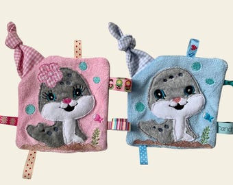 May include: Two plush baby toys with embroidered seal faces. The seal on the left is pink with a flower and the seal on the right is blue. Both toys have multiple ribbons attached.