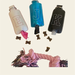 Könnte beinhalten: Drei personalisierte Hundekotbeutelhalter in Weiß, Blau und Schwarz, jeweils mit einem Buchstaben. Rosa Hundekotbeutel und braune knochenförmige Leckerlis sind verstreut. Ein rosa Hundespielzeug aus Seil befindet sich unten.