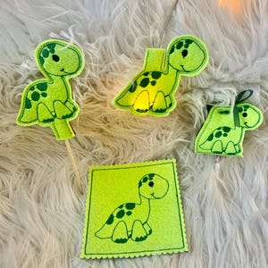 Könnte beinhalten: Vier grüne Filz-Dinosaurier-Ornamente mit schwarzen Punkten. Ein Dinosaurier ist auf einem Stock, einer auf einer Schnur, einer auf einem Quadrat und einer auf einer Schnur mit einer Schleife.