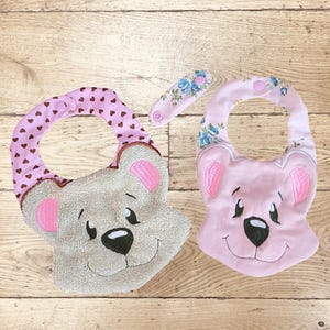 May include: Two baby bibs in the shape of teddy bears. One is light grey with pink ears and a brown heart-patterned neckband. The other is pink with blue floral accents and pink ears. Both have smiling faces and black eyes.