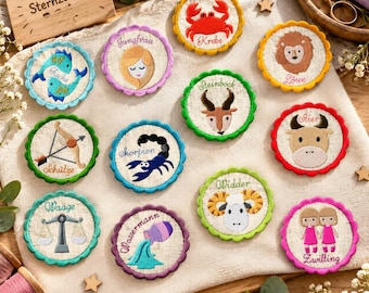 May include: A collection of twelve circular embroidered patches featuring zodiac signs with German text labels. Each patch has a scalloped edge and depicts a unique astrological symbol, including animals and figures, arranged on a light fabric surface.