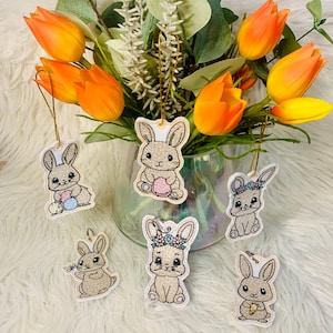Könnte beinhalten: Sechs bestickte Hasen-Anhänger mit verschiedenen Designs. Jeder Anhänger zeigt einen Hasen mit einem anderen Ausdruck und hält ein anderes Objekt, wie ein Ei, eine Karotte oder eine Blume. Die Anhänger sind alle weiß mit goldfarbener Stickerei.