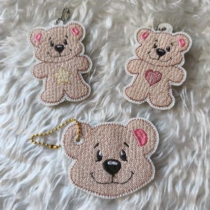 Könnte beinhalten: Drei bestickte Teddybär-Schlüsselanhänger. Die Bären sind hellbraun mit rosa Akzenten an den Ohren und einer schwarzen Nase. Ein Bär hat ein Herz, ein anderer einen Stern und der dritte ein lächelndes Gesicht. Jeder Schlüsselanhänger hat eine Metallkette.