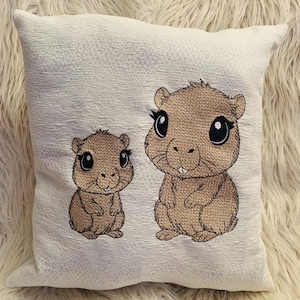 Könnte beinhalten: Ein weißes Kissen mit gestickten Bildern von zwei braunen Hamstern mit großen Augen. Der größere Hamster lächelt und der kleinere Hamster schaut geradeaus.