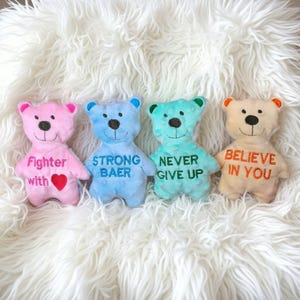 Könnte beinhalten: Vier farbenfrohe Plüschteddys mit gestickten Gesichtern und inspirierenden Botschaften. Die Bären sind rosa, blau, türkis und beige, mit Texten wie "Fighter with", "Strong Baer", "Never Give Up" und "Believe in You".