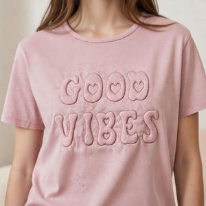 Könnte beinhalten: Ein hellrosa T-Shirt mit kurzen Ärmeln und dem aufgestickten Schriftzug "GOOD VIBES" in einer erhabenen, blasenartigen Schriftart. Das Wort "GOOD" hat herzförmige Akzente. Das Shirt ist aus weichem Material.