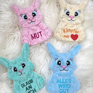 Könnte beinhalten: Vier Plüsch-Hasenspielzeuge in Pastellfarben: Rosa, Gelb, Türkis und Blau. Jeder Hase hat gestickte Gesichtszüge, Wimpern und Text auf dem Bauch. Der rosa Hase sagt "MUT", der gelbe Hase sagt "Kämpfer mit", der türkisfarbene Hase sagt "GLAUB AN DICH" und der blaue Hase sagt "ALLES WIRD GUT".