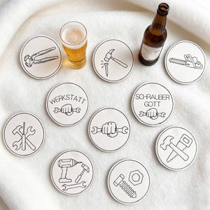 May include: A collection of round, off-white coasters with black line drawings of tools and text. Tools include pliers, a hammer, a nail, a saw, a wrench, a screwdriver, a drill, and a bolt. A glass of beer and a brown bottle are also present.