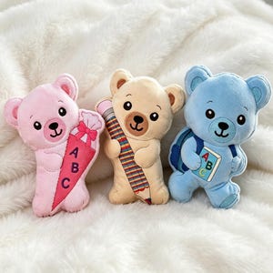 Puede incluir: Tres osos de peluche en rosa, beige y azul. El oso rosa sostiene un cono con un lazo rosa y las letras ABC. El oso beige sostiene un lápiz grande multicolor. El oso azul lleva una mochila azul y sostiene un libro con las letras ABC.