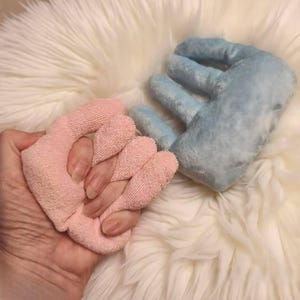 以下が含まれることがあります： ピンク色の指用手袋をつけた手と、青いぬいぐるみのような手のクローズアップ。ピンクの手袋は柔らかく、質感のある素材で作られています。青いぬいぐるみのような手はふわふわで柔らかいです。背景は白くふわふわした表面です。