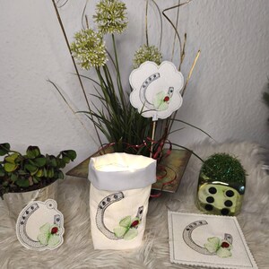 May include: A collection of decorative items featuring a horseshoe and four-leaf clover design. Includes a small fabric bag, a coaster, a tag, and a decorative pick. The items are embroidered with silver thread, green clovers, and red accents.