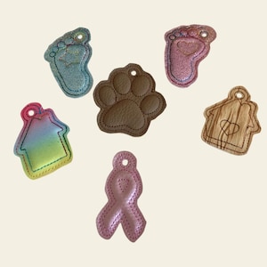 May include: A collection of embroidered charms in various shapes, including a baby footprint, a paw print, a house, and a pink ribbon. The charms are in shades of blue, pink, brown, and rainbow colors, with a stitched outline and a hole for hanging.