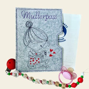 Pode incluir: Capa de feltro cinza com a palavra "Mutterpass" bordada em roxo. A capa apresenta uma ilustração de desenho animado de uma mulher grávida segurando uma meia de bebê. Há quatro corações vermelhos bordados acima da mulher.
