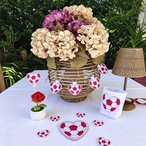 May include: A decorative arrangement featuring a woven basket filled with beige and purple hydrangeas. A string of heart-shaped soccer ball decorations hangs from the basket. Additional soccer ball hearts and a small white pot with a red rose are on the white tablecloth.