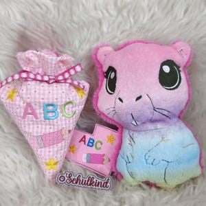 May include: A pink and purple plush animal with large black eyes and a small, pink, heart-shaped ABC card. A pink gingham bag with the letters ABC and a pencil design is also present. The word "Schulkind" is on a small label.