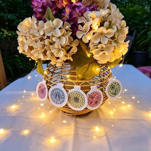 May include: A gold wire basket holds a floral arrangement of beige and purple flowers, illuminated by fairy lights. Decorative, embroidered ornaments with floral designs hang from the basket, adding a touch of whimsy to the display.