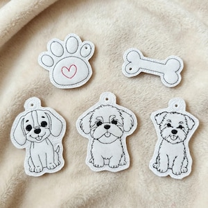 Puede incluir: Un conjunto de cinco parches bordados blancos que presentan tres ilustraciones de perros distintas, una huella con un corazón rojo cosido y una forma de hueso. Cada parche tiene un pequeño orificio en la parte superior, con contornos negros.