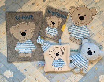 May include: A collection of baby items featuring a teddy bear design. Includes a gray felt cover with "U-Heft" text, a small "Impfpass" cover, a plush bear, and a soft toy, all with blue and white striped shirts.