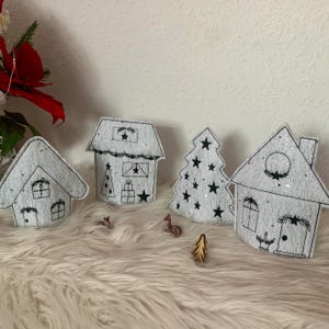 Puede incluir: Un conjunto de cuatro decoraciones navideñas blancas, que incluyen tres casas y un árbol de Navidad. Las decoraciones están hechas de fieltro y tienen contornos negros. Las casas tienen ventanas y puertas, y el árbol de Navidad tiene estrellas.