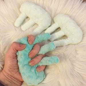 May include: Three plush hand-shaped toys in the image. One is held by a hand, the other two are next to it. The held toy is a teal color, and the other two are a light green. All three have a textured, bumpy surface.