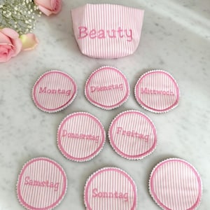 May include: A pink and white striped cosmetic bag with the word "Beauty" embroidered on it. Seven round, pink and white striped fabric circles with the days of the week embroidered on them.