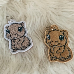 May include: Two embroidered keychains featuring cartoon brown hamsters with big eyes. The hamsters are standing on their hind legs with their front paws in front of them. The keychains are made of felt and have a white background.