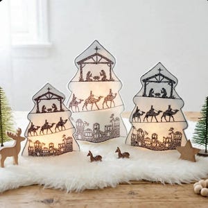 May include: Three white, tree-shaped, illuminated decorative structures featuring black silhouette illustrations of a nativity scene, camels, and a town. The set includes small wooden deer figures and a star, arranged on a white faux fur base.